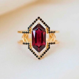 May include: A gold-toned ring featuring a large, rectangular, deep red gemstone at its center. The gemstone is framed by a black beaded border within a geometric design. Additional black beads and gold accents complete the ring's design.