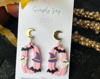 May include: Pink Halloween-themed earrings with gold crescent moon accents. Each earring features a pink pumpkin shape with a black bat, a purple witch hat, and a white ghost. The earrings are from Simply Zay.