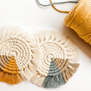 Op de afbeelding: Twee handgemaakte macramé onderzetters met een cirkelvormig ontwerp. De onderzetters hebben een crème, bruin, mosterdgeel en grijze kleurstelling. Een spoel gele macramé draad staat op de achtergrond. De onderzetters hebben een gefranjerde rand.