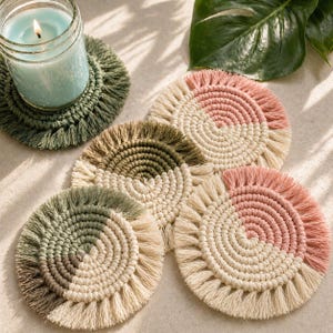 May include: A collection of handmade macrame coasters in various colors, including green, pink, and cream. The coasters have a circular design with a fringed edge, perfect for protecting surfaces from drink condensation. A lit candle in a glass jar sits on one of the coasters.