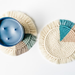 May include: Two round macrame coasters with fringe. One coaster has a blue candle on it. The other coaster has a teal, tan, and cream color block design. The coasters are on a white background.