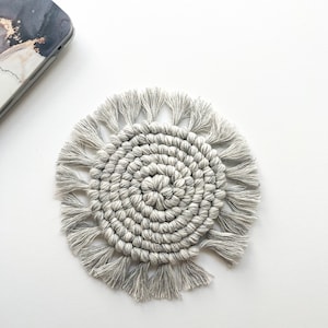 Op de afbeelding: Een grijze macramé onderzetter met een spiraalvormig ontwerp en franjes. De ronde onderzetter ligt op een witte ondergrond. Een telefoonhoesje met marmerpatroon is zichtbaar.