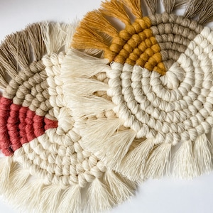 Op de afbeelding: Set van drie handgemaakte macramé onderzetters. Elke onderzetter heeft een cirkelvormig ontwerp met crème, mosterdgeel, rood en bruin. De onderzetters hebben een gefranjerde rand.