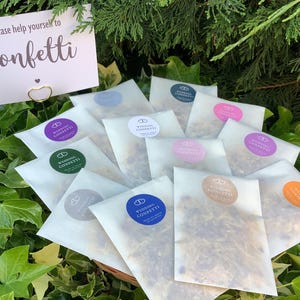 Könnte beinhalten: Kleine, weiße Päckchen mit Konfetti, jedes mit einem farbigen Aufkleber verschlossen. Ein Schild mit der Aufschrift "Please help yourself to Confetti" steht davor. Die Päckchen sind auf grünem Laub angeordnet.