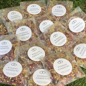 May include: Clear cellophane bags filled with colorful wedding confetti. Each bag has a white circular sticker with the words "WEDDING CONFETTI" and "Thanks for sharing our special day". The confetti is a mix of dried petals.