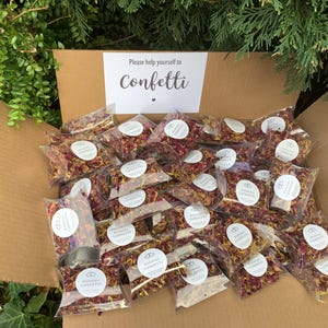 May include: A cardboard box filled with numerous small, clear plastic bags containing dried flower petals in shades of pink, yellow, and orange. Each bag features a circular white label with the text Wedding Confetti. A sign inside reads Please help yourself to Confetti.