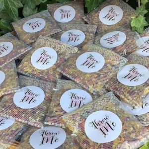 May include: A selection of small mesh bags filled with dried flower petals. Each bag features a white circular sticker with the words "Throw Me" in elegant script, with floral accents. The bags are arranged in a woven basket.