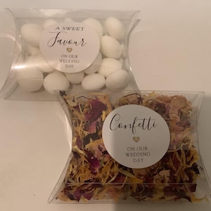 May include: Two clear pillow-shaped boxes containing wedding favours. The top box holds white sweets with a label reading A SWEET Favour ON OUR WEDDING DAY. The bottom box contains dried flower petals with a label reading Confetti ON OUR WEDDING DAY.