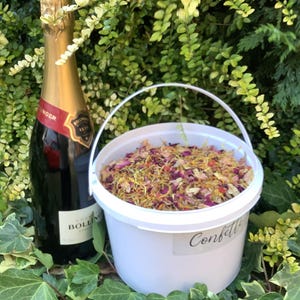 May include: A white plastic bucket filled with a mixture of dried flower petals in shades of pink, yellow, and orange, featuring a label with the text Confetti, positioned next to a dark glass bottle of Bollinger champagne with a gold foil neck.