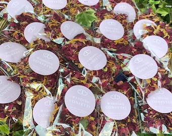 May include: Multiple small clear plastic bags filled with red and yellow flower petals. Each bag has a round pink label with the text "Wedding Confetti" printed in black.