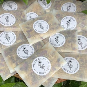 Op de afbeelding: Een verzameling kleine, rechthoekige pakketjes gevuld met gedroogde bloemenconfetti. Elk pakketje is verzegeld met een witte sticker met de tekst "Just Married" en een bloemenillustratie. De confetti is een mix van gele, rode en bruine bloemblaadjes.