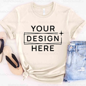 May include: A beige t-shirt with short sleeves and a crew neck. The shirt has black text that says "YOUR DESIGN HERE" in a box.