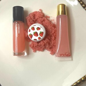 May include: A collection of beauty products, including a lip gloss tube with a black cap, a lip gloss tube with a gold cap, and a small container with a strawberry design. Pink scrub is in the center. The products are arranged on a white plate with a gold rim.