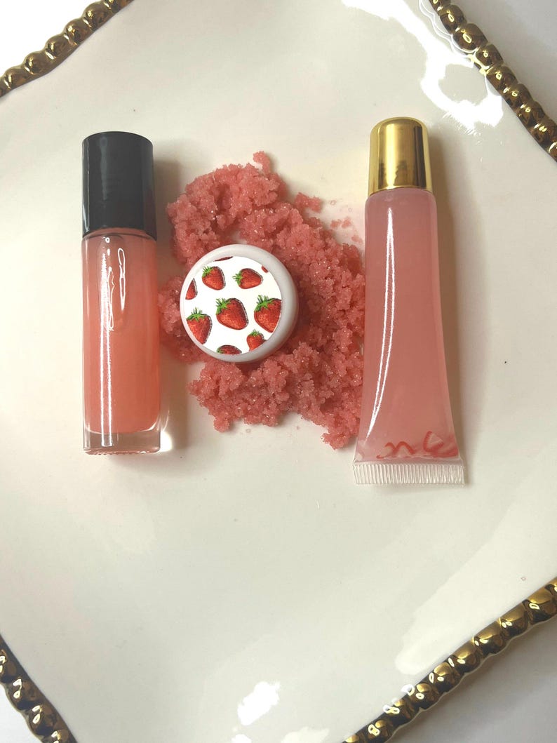May include: A collection of beauty products, including a lip gloss tube with a black cap, a lip gloss tube with a gold cap, and a small container with a strawberry design. Pink scrub is in the center. The products are arranged on a white plate with a gold rim.