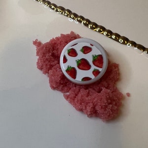 May include: A white, round container lid with a strawberry pattern sits on a pile of pink, granular material. A gold-colored chain is in the background. The strawberries are red with green leaves.