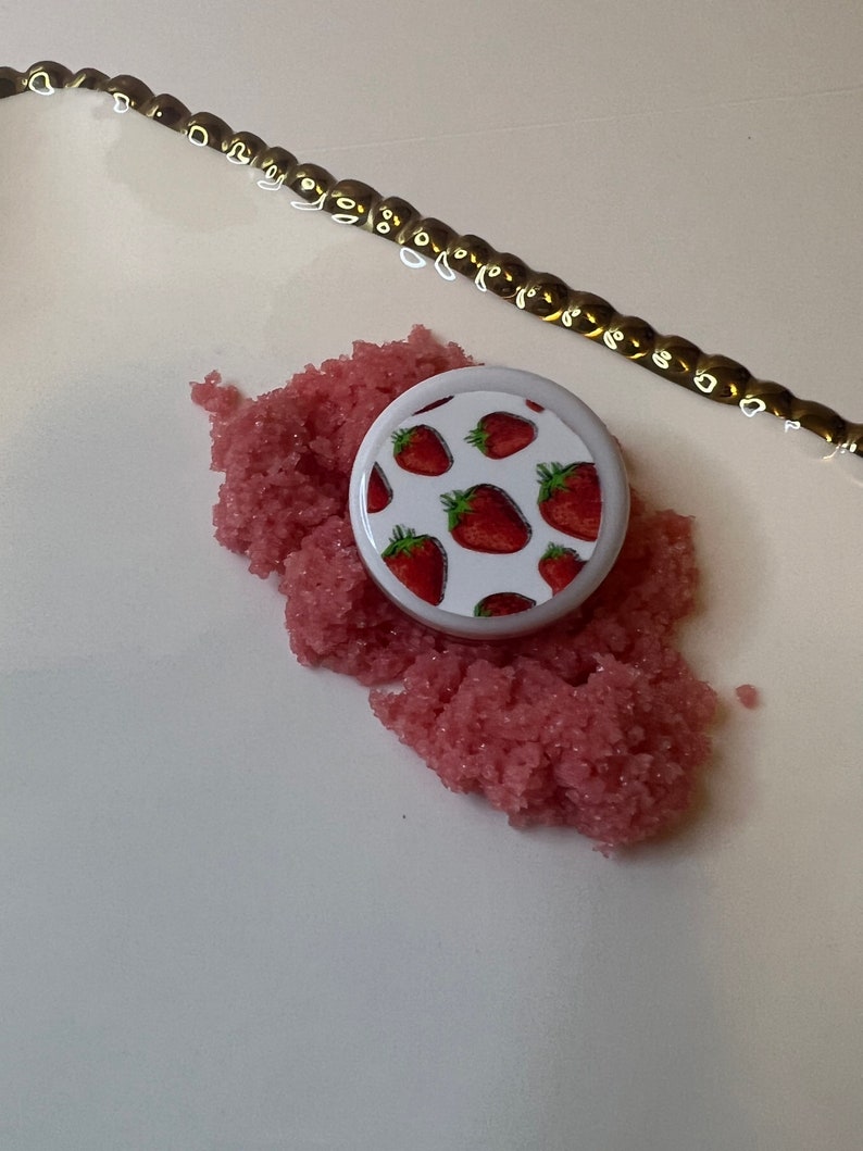 May include: A white, round container lid with a strawberry pattern sits on a pile of pink, granular material. A gold-colored chain is in the background. The strawberries are red with green leaves.