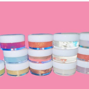 May include: A collection of 12 small jars filled with colorful lip gloss. The jars are clear with white lids and the lip glosses are various shades of pink, orange, blue, green, and purple.