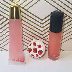 May include: Three lip gloss products. Two pink lip gloss tubes with gold and black caps, and a small round container with a strawberry pattern on the lid. The background is white and gold.