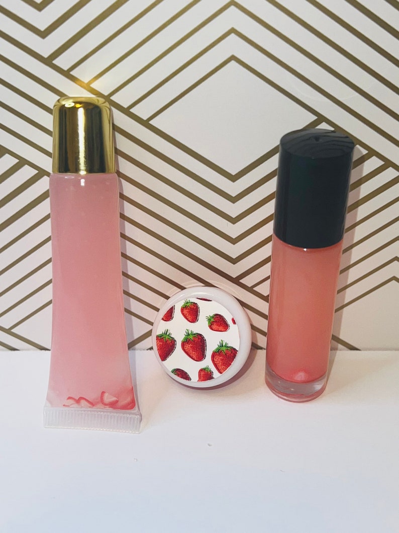 May include: Three lip gloss products. Two pink lip gloss tubes with gold and black caps, and a small round container with a strawberry pattern on the lid. The background is white and gold.
