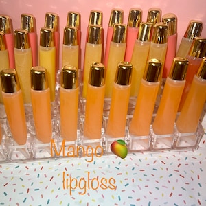 May include: A clear plastic container filled with 24 small tubes of mango lip gloss. The tubes have gold lids and the lip gloss is a light orange color. The container is sitting on a white surface with a colorful confetti pattern. The text "Mango lip gloss" is written below the container.