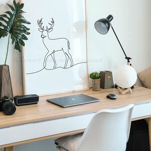 May include: A framed black and white line drawing of a deer on a white background. The drawing is on a desk with a laptop, a lamp, and a moon lamp. The desk is made of wood and has a white drawer. A white chair with wooden legs is in the foreground.