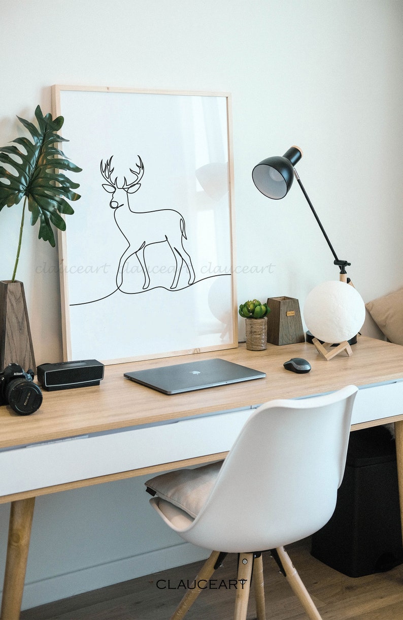 May include: A framed black and white line drawing of a deer on a white background. The drawing is on a desk with a laptop, a lamp, and a moon lamp. The desk is made of wood and has a white drawer. A white chair with wooden legs is in the foreground.