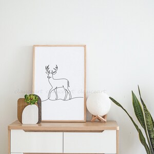 May include: A framed black and white line drawing of a deer in a minimalist style. The frame is made of wood and has a light brown finish. The drawing is on a white background. The frame is sitting on a white dresser with two drawers. There is a small potted plant and a white moon lamp on the dresser. The dresser is against a white wall.