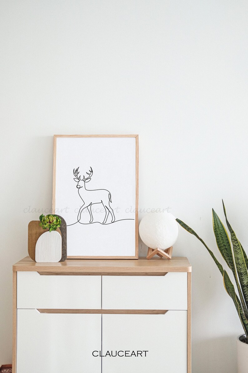 May include: A framed black and white line drawing of a deer in a minimalist style. The frame is made of wood and has a light brown finish. The drawing is on a white background. The frame is sitting on a white dresser with two drawers. There is a small potted plant and a white moon lamp on the dresser. The dresser is against a white wall.