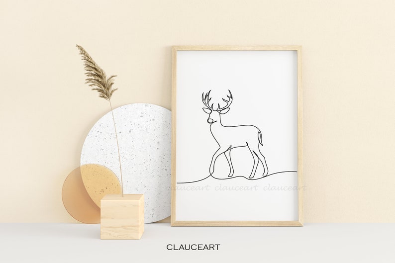 May include: A black and white line drawing of a deer with antlers, framed in a light wood frame. The frame is sitting on a white surface with a beige background.