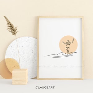 Pu&ograve; includere: Una stampa minimalista di arte lineare di una persona che fa surf su un'onda con un sole color pesca sullo sfondo. La stampa &egrave; incorniciata in una cornice di legno chiaro ed &egrave; esposta su una superficie bianca.