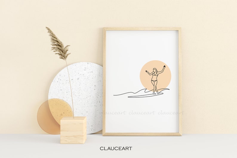 Pu&ograve; includere: Una stampa minimalista di arte lineare di una persona che fa surf su un'onda con un sole color pesca sullo sfondo. La stampa &egrave; incorniciata in una cornice di legno chiaro ed &egrave; esposta su una superficie bianca.