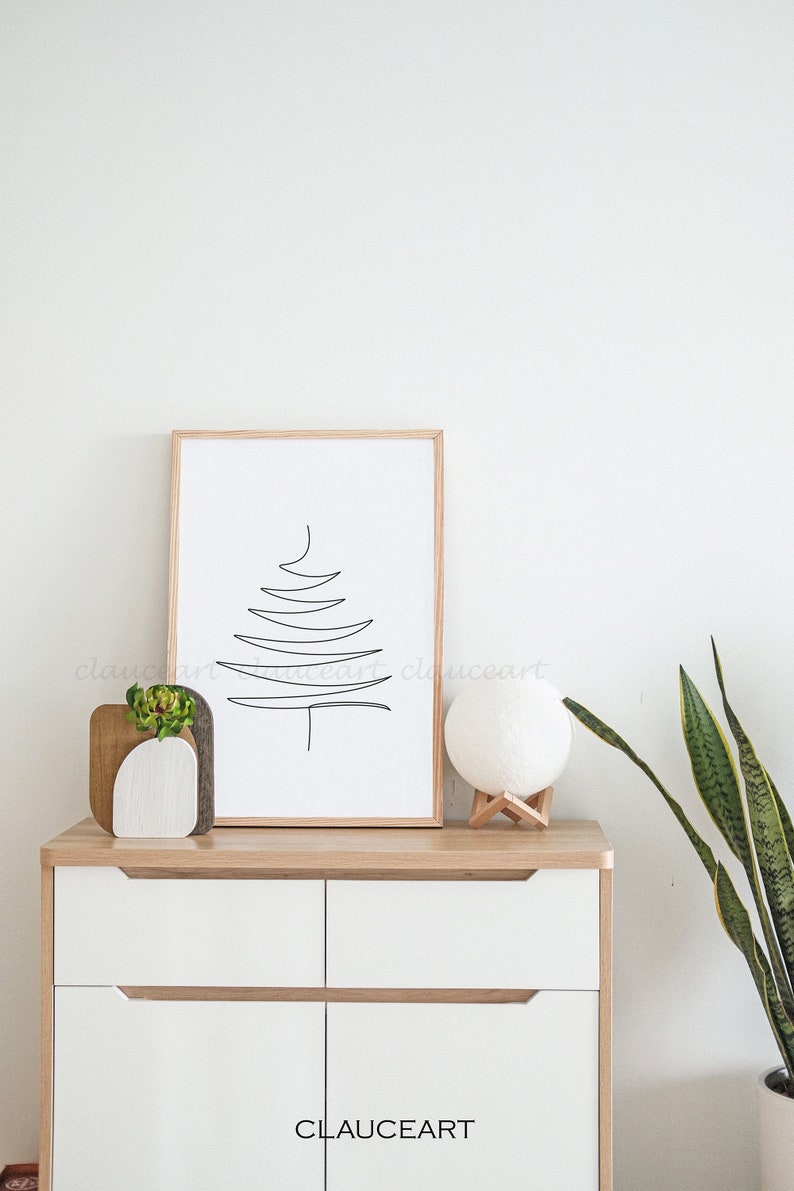 May include: A framed black and white line drawing of a stylized Christmas tree on a white background. The frame is made of light wood. The drawing is on a wooden shelf with a white ceramic vase with a small green plant, a wooden decorative object, and a white moon lamp on a wooden stand. The shelf is part of a white cabinet with two drawers. The cabinet has a wooden top and wooden drawer pulls. A green snake plant is in a white pot on the right side of the image.