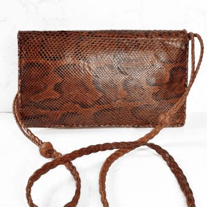 May include: A brown leather crossbody bag with a snakeskin pattern. The bag has a rectangular shape and a braided strap. The bag is made of a textured material and has a flap closure. The bag is a neutral colour and would be suitable for everyday use.