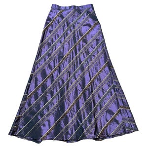 Pode incluir: Uma saia longa e fluida em um tom roxo profundo, com listras diagonais adornadas com detalhes dourados e marrons. A saia tem um leve brilho, sugerindo um tecido acetinado ou semelhante à seda. O design é elegante e adequado para ocasiões formais.