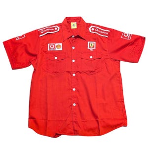 Könnte beinhalten: Rotes kurzärmeliges Hemd mit mehreren Logos, darunter "Vodafone" und dem Ferrari-Emblem. Das Hemd hat zwei Brusttaschen und weiße Knöpfe. Das Ferrari-Logo befindet sich auf der Brust. Das Hemd hat eine leuchtend rote Farbe.