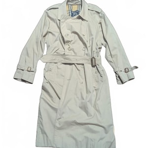 Op de afbeelding: Een beige trenchcoat met een dubbele rij knopen, met een riem in de taille en bandjes op de mouwen. De jas heeft een klassieke kraag en een geruite voering zichtbaar bij de nek. De jas is gemaakt van een lichtgewicht materiaal.