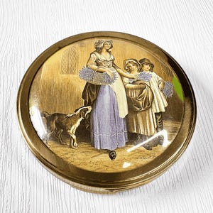 Könnte beinhalten: Ein runder, antiker goldfarbener Kompaktpuder mit einer gemalten Szene einer Frau und zwei Kindern, die Körbe mit Lavendel tragen, mit einem Hund. Der Kompaktpuder hat eine glänzende Oberfläche und ein detailliertes Vintage-Design.