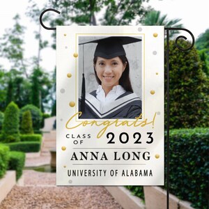 May include: A white graduation garden flag with gold and silver confetti. The flag features a photo of a graduate wearing a cap and gown, the text "Congrats! Class of 2023 Anna Long University of Alabama", and a white background.