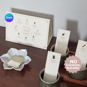 May include: A cream-colored table tent with the text "What will Leah choose?" and various icon options. Small cards with icons and text are in ceramic cups. A white flower-shaped dish holds business cards.
