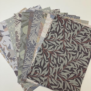 May include: A collection of patterned papers fanned out, showcasing various botanical and floral designs. The papers feature a range of colors, including muted greens, blues, and browns, with intricate leaf and flower motifs. The papers are arranged on a white surface.