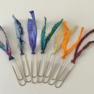 May include: Eight silver paper clips with colorful fabric strips attached. The fabric strips are in shades of blue, green, yellow, orange, and burgundy.