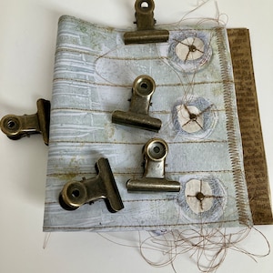 May include: A close-up of a handmade journal with five antique brass binder clips holding the pages together. The journal has a white background with a light green and brown pattern. The pages are stitched together with brown thread. The journal is on a white surface.