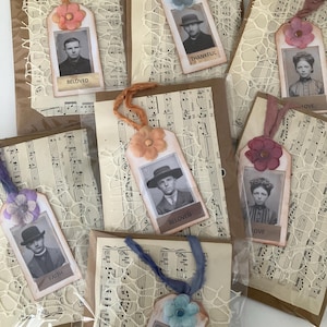 May include: A collection of seven rectangular gift tags featuring vintage black and white portrait photographs, each adorned with a fabric flower, a coloured ribbon tie, and a decorative lace overlay, all mounted on sheets of printed musical notation.