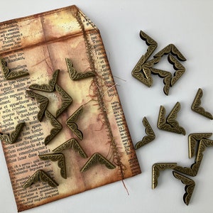 May include: A collection of antique brass corner protectors with a decorative design. The corner protectors are arranged on a piece of paper with text.