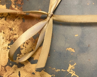 Könnte beinhalten: Nahaufnahme eines cremefarbenen Satinbands, das zu einer Schleife gebunden ist. Das Band liegt auf einer Oberfläche mit einem blauen und goldenen Farbschema. Die Schleife ist mittig platziert, das Band erstreckt sich nach außen.
