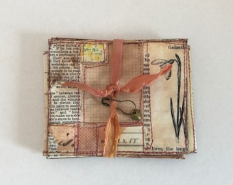 May include: A handmade, vintage-style journal with pages of printed text and grid patterns. It is bound with a pink ribbon and secured with a safety pin and a heart-shaped charm. The cover features a floral illustration and the word "Galant".