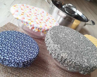 Puede incluir: Una colección de cubiertas de tela redondas para cuencos con bordes elásticos, con un estampado de cachemira azul, un patrón floral gris y un diseño geométrico de triángulos multicolor, junto a una olla de acero inoxidable con tapa.