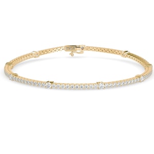 May include: A delicate gold tennis bracelet with numerous small, sparkling white gemstones set along its length. The bracelet features a secure clasp and is displayed against a white background. The brand name "Skosh" is visible.