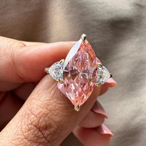 May include: A gold ring featuring a large, pink, marquise-cut gemstone as the centerpiece, flanked by two smaller, pear-shaped, white gemstones. The ring is held up, showcasing the intricate facets of the stones.