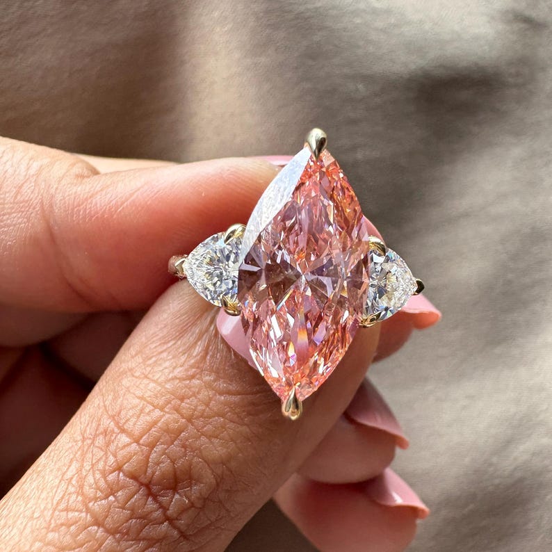 May include: A gold ring featuring a large, pink, marquise-cut gemstone as the centerpiece, flanked by two smaller, pear-shaped, white gemstones. The ring is held up, showcasing the intricate facets of the stones.